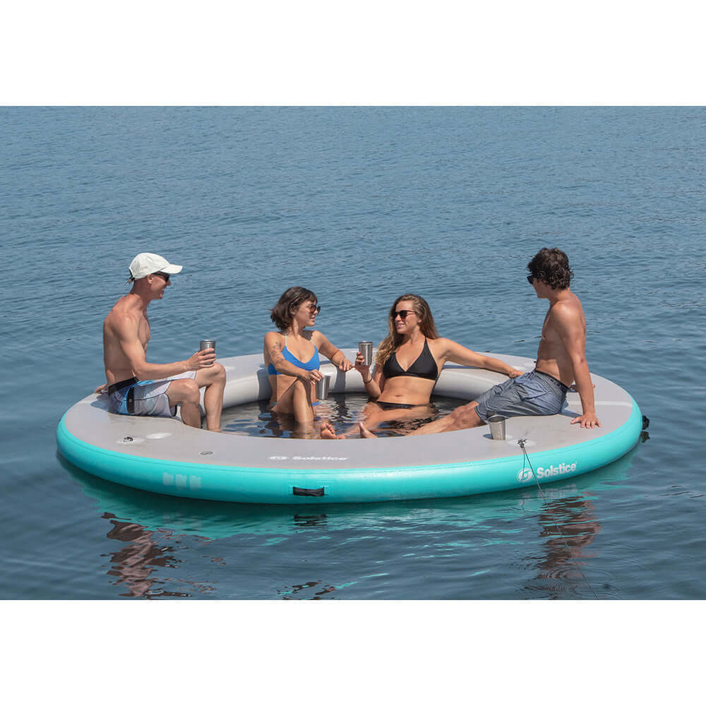 Four people relaxing on a Solstice Watersports circular mesh dock in the middle of a lake.