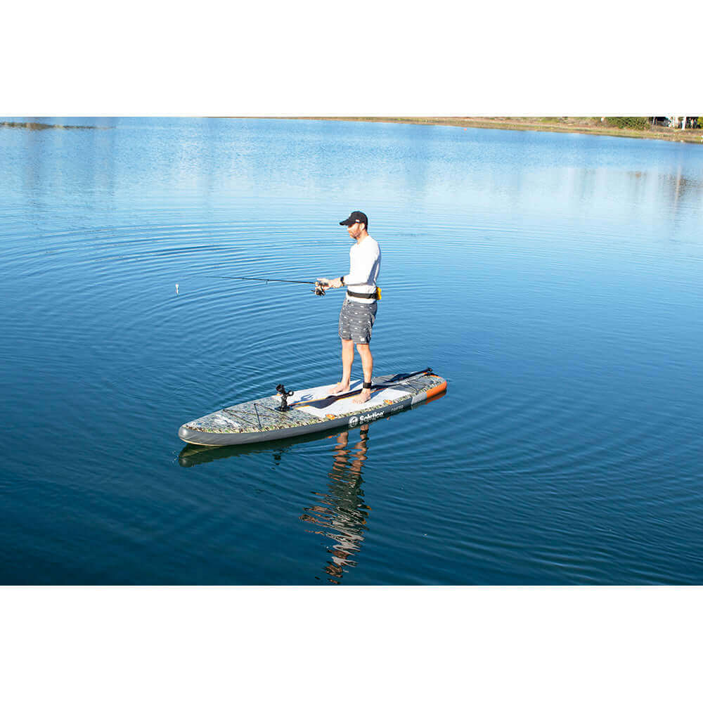 Person fishing on Solstice Watersports inflatable paddleboard in serene lake setting.