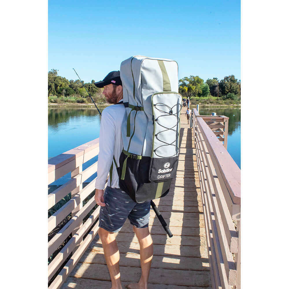 Man carrying Solstice Drifter paddleboard kit on a backpack along a lakeside pier.