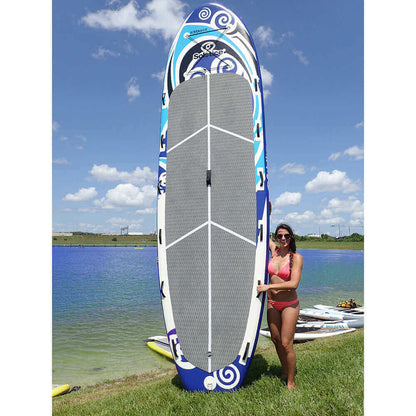 Woman standing beside Solstice Watersports 16' Maori Giant Inflatable Paddleboard near a lake.