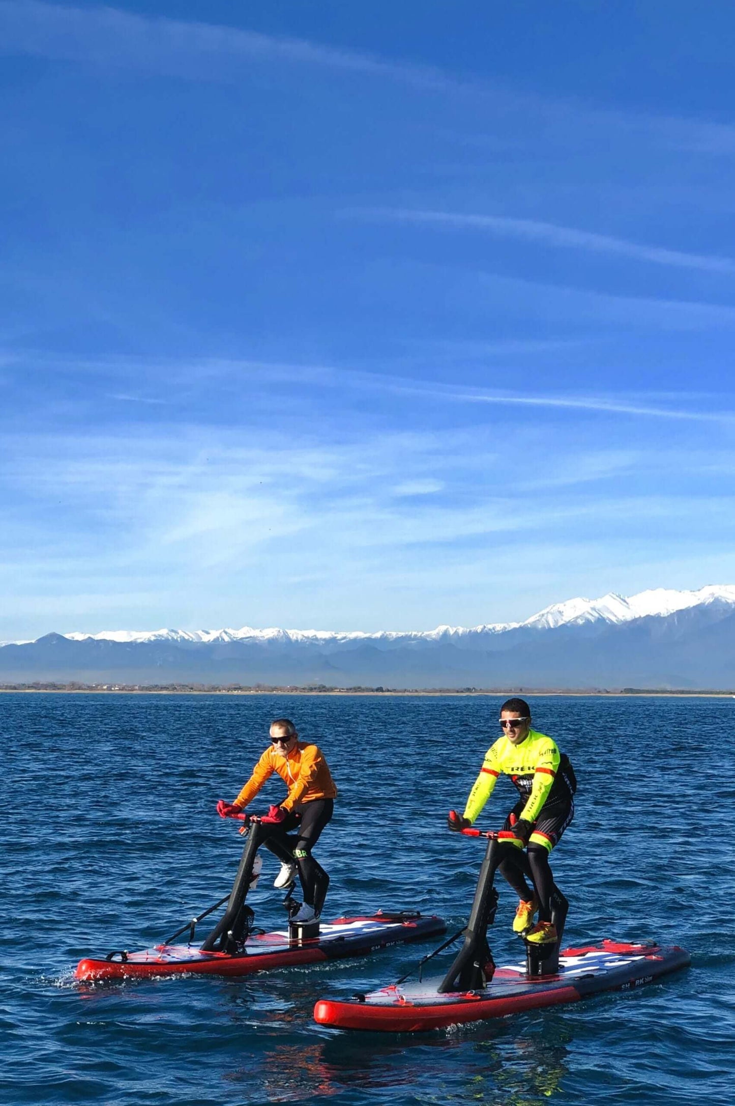 Two people enjoying a workout on Red Shark fitness boards on a clear blue lake with mountains in the background.