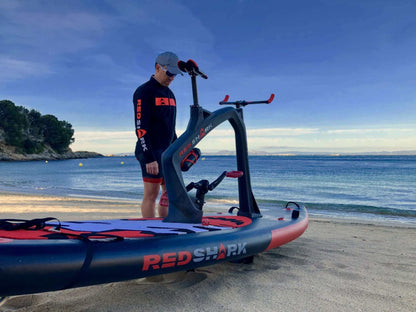 Man standing next to Red Shark fitness water craft on a sandy beach, ready for a workout on the ocean.